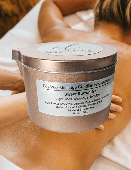 6oz CandleLux Sweet Surrender luxury massage candle in a black travel candle tin with pour spout and a picture of a woman getting a massage in the background.